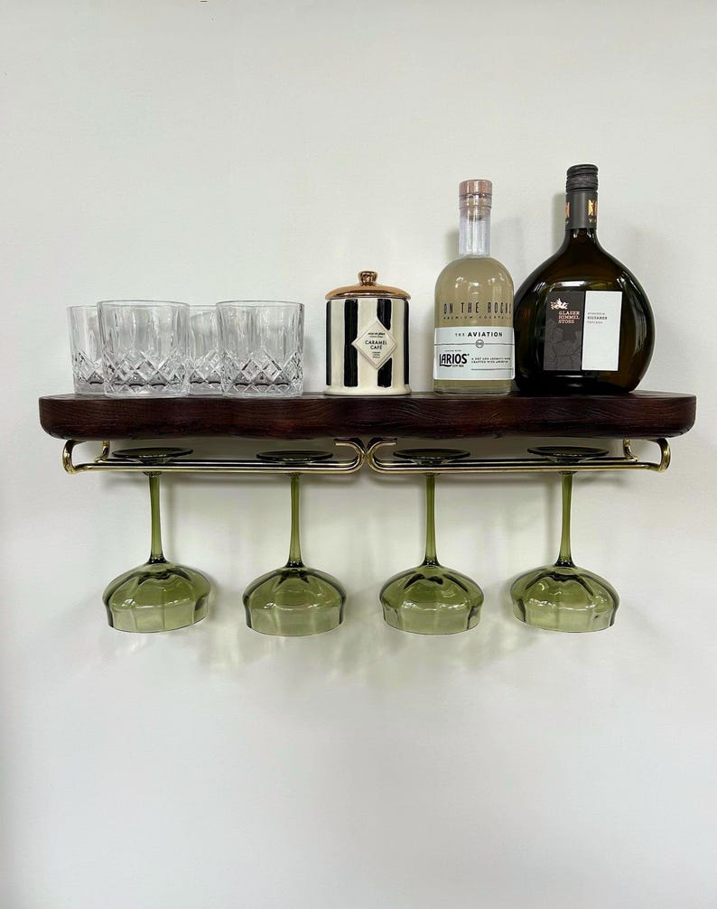 May include: A dark wood wall-mounted shelf with a gold metal rack holding four green-tinted stemmed glasses. The shelf top displays three clear crystal tumblers, a black and white striped canister with a gold lid, and two glass bottles.