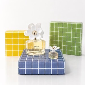 May include: Two perfume bottles, one large and one small, sit on a blue tiled surface. The larger bottle has a white daisy flower cap and the text "DAISY MARC JACOBS". A yellow and green tiled block are behind the perfume.