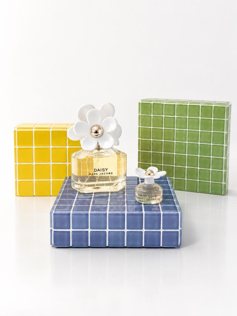 May include: Two perfume bottles, one large and one small, sit on a blue tiled surface. The larger bottle has a white daisy flower cap and the text "DAISY MARC JACOBS". A yellow and green tiled block are behind the perfume.
