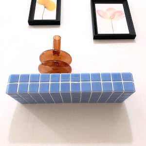 Puede incluir: Una estanter&iacute;a de azulejos azules con un jarr&oacute;n de vidrio marr&oacute;n en la parte superior. El jarr&oacute;n est&aacute; apilado con dos platos redondos de vidrio marr&oacute;n. Dos cuadros enmarcados con estampados florales est&aacute;n en la pared por encima de la estanter&iacute;a.