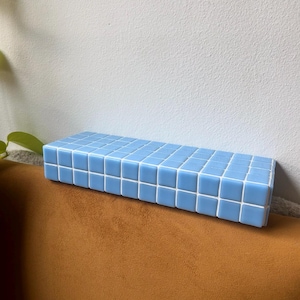 Puede incluir: Una estanter&iacute;a de color azul claro hecha de peque&ntilde;as baldosas cuadradas. La estanter&iacute;a est&aacute; montada en una pared y est&aacute; vac&iacute;a.
