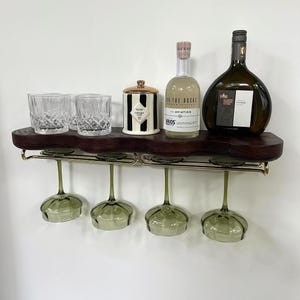 May include: A dark wood wall-mounted shelf with a live edge, featuring a gold metal rack holding four green-tinted stemmed glasses. On the shelf sit three clear crystal tumblers, a black and white striped jar, and two glass bottles of alcohol.