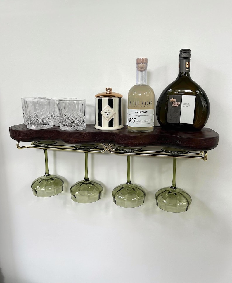 May include: A dark wood wall-mounted shelf with a live edge, featuring a gold metal rack holding four green-tinted stemmed glasses. On the shelf sit three clear crystal tumblers, a black and white striped jar, and two glass bottles of alcohol.