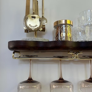 May include: A dark wood wall-mounted shelf with a gold metal rack holding three stemmed glasses. On the shelf sits a gold bar tool set on a stone base, a clear jar with a gold lid labeled DEHY, and two clear textured glass tumblers.
