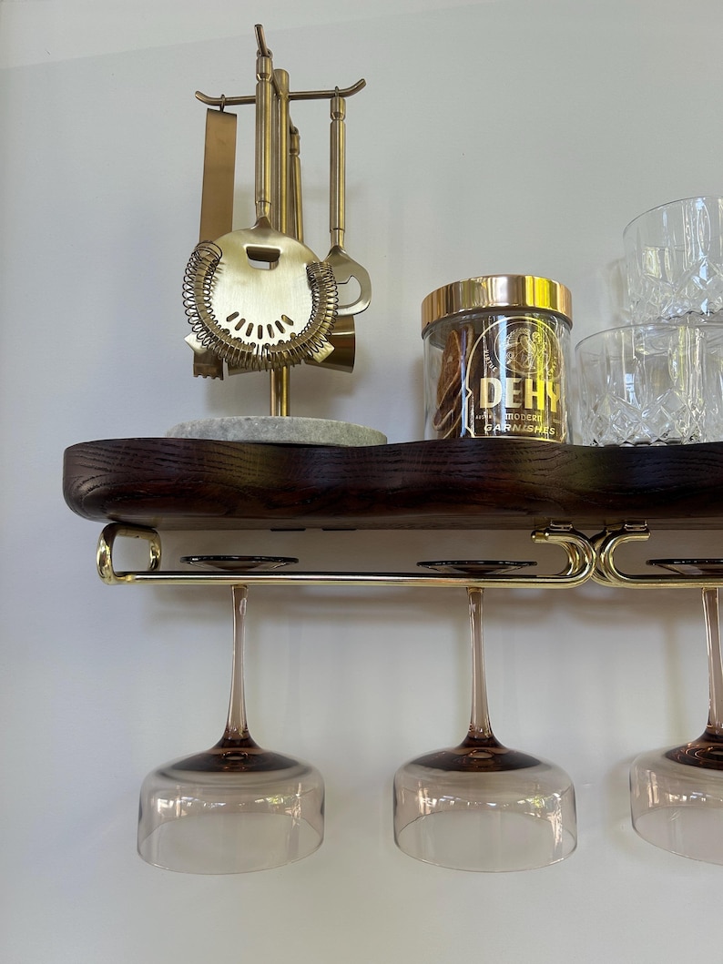 May include: A dark wood wall-mounted shelf with a gold metal rack holding three stemmed glasses. On the shelf sits a gold bar tool set on a stone base, a clear jar with a gold lid labeled DEHY, and two clear textured glass tumblers.