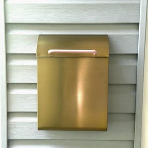 May include: A rectangular wall-mounted mailbox with a brushed gold metallic finish and a smooth, light pink horizontal handle attached to the top lid. The mailbox features a clean, vertical design with a curved top flap for opening and closing.