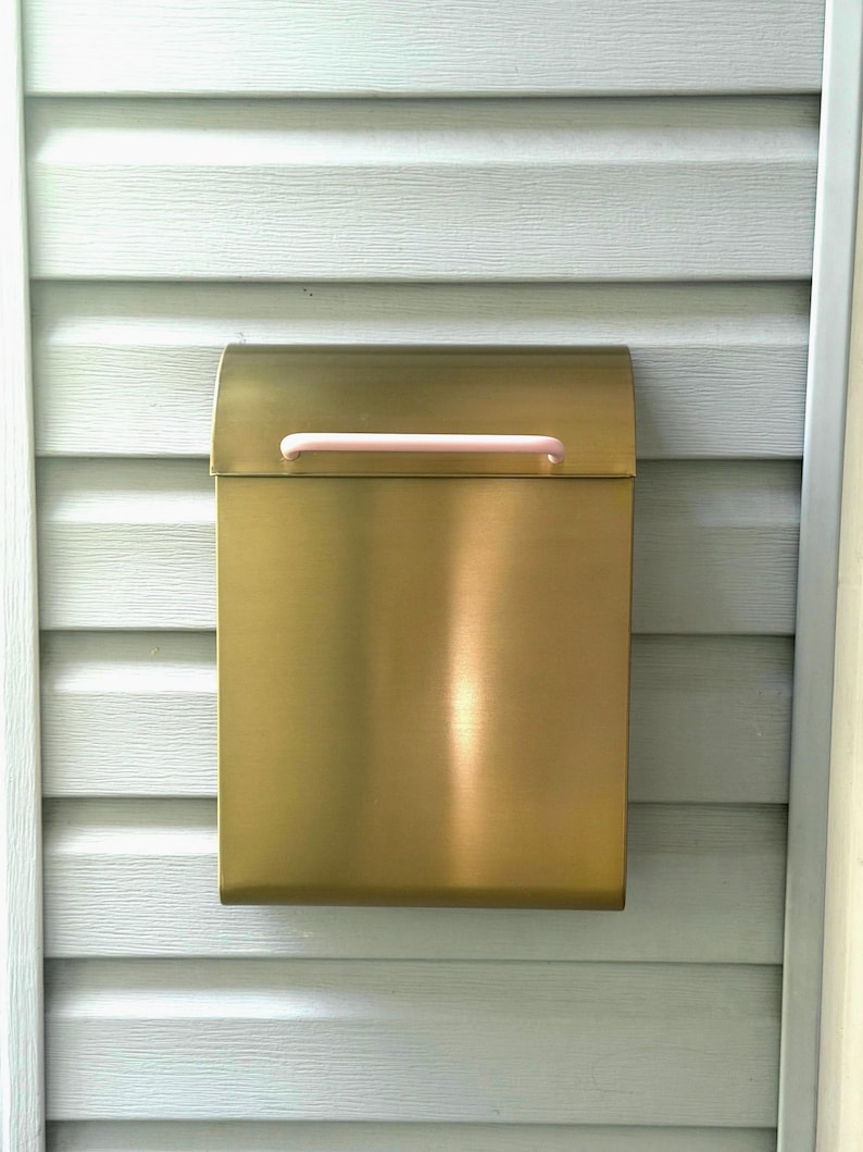 May include: A rectangular wall-mounted mailbox with a brushed gold metallic finish and a smooth, light pink horizontal handle attached to the top lid. The mailbox features a clean, vertical design with a curved top flap for opening and closing.