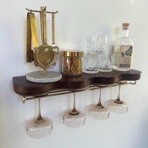 May include: A dark wood wall-mounted shelf with a wavy front edge and gold metal hardware. It holds a gold bar tool set on a marble base, a glass jar of dried citrus, three crystal tumblers, a gin bottle, and four hanging stemmed glasses.