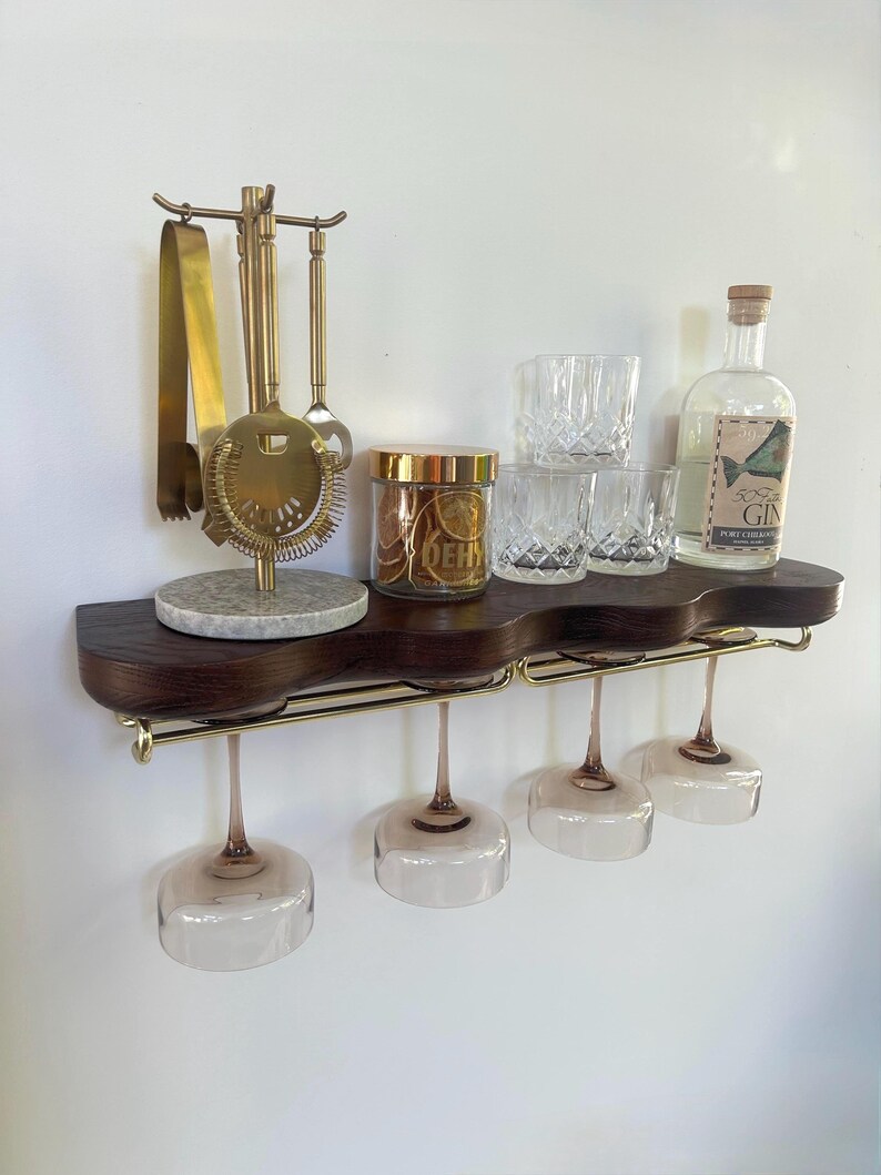 May include: A dark wood wall-mounted shelf with a wavy front edge and gold metal hardware. It holds a gold bar tool set on a marble base, a glass jar of dried citrus, three crystal tumblers, a gin bottle, and four hanging stemmed glasses.