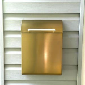 May include: A rectangular wall-mounted mailbox with a brushed gold metallic finish. The mailbox features a curved top lid and a horizontal white handle centered on the front flap for opening. The design is simple, clean, and functional.