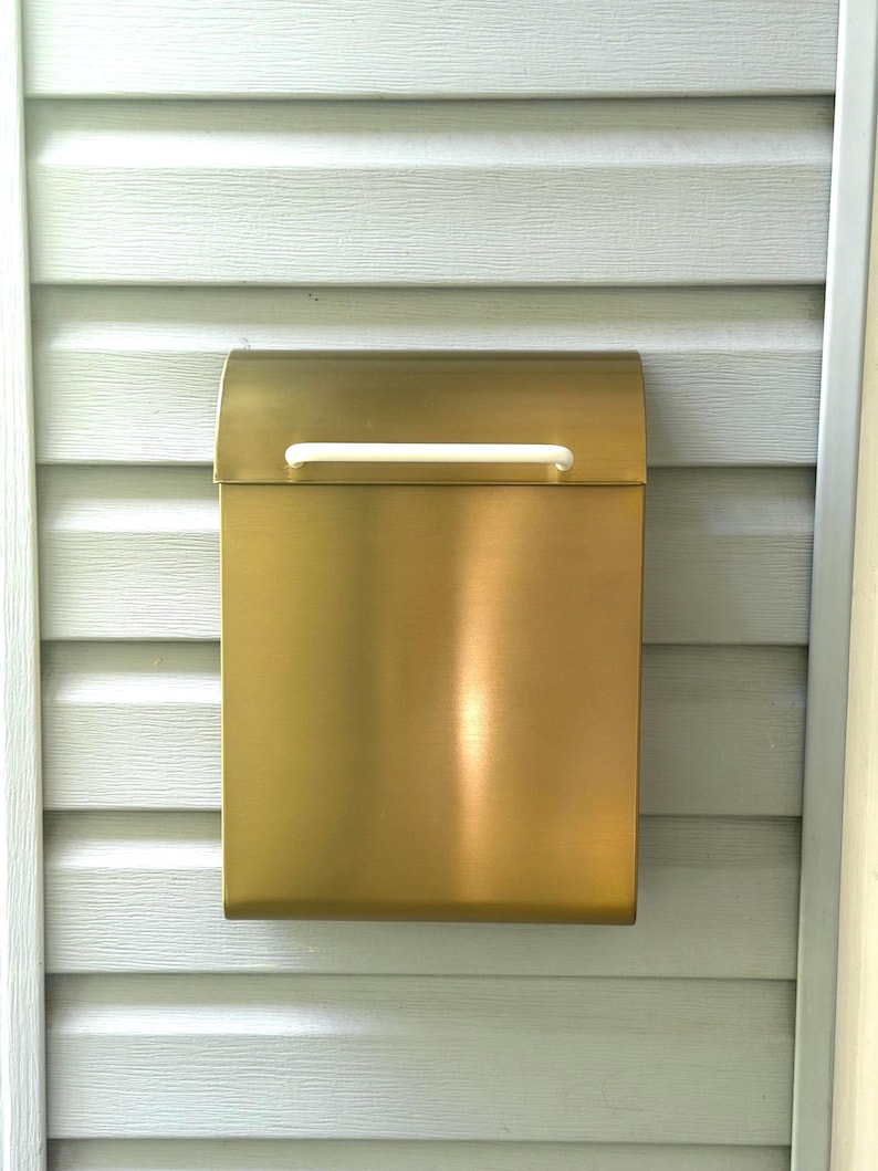 May include: A rectangular wall-mounted mailbox with a brushed gold metallic finish. The mailbox features a curved top lid and a horizontal white handle centered on the front flap for opening. The design is simple, clean, and functional.