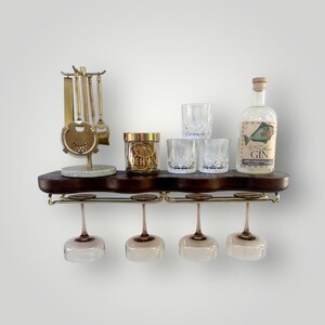May include: A dark wood wall-mounted bar shelf featuring a gold-toned metal rack for four hanging stemware glasses. The shelf holds a gold bar tool set, a small glass jar, two clear textured tumblers, and a bottle of 50 Fathoms Gin with a fish label.