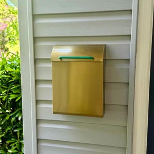 May include: A rectangular wall-mounted mailbox made of brushed brass with a smooth, metallic finish. The mailbox features a curved top lid and a thin, horizontal green handle centered on the front flap for opening and closing the compartment.