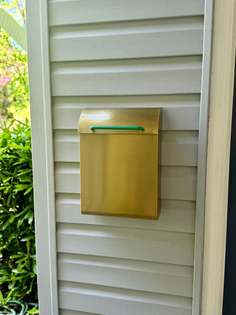 May include: A rectangular wall-mounted mailbox made of brushed brass with a smooth, metallic finish. The mailbox features a curved top lid and a thin, horizontal green handle centered on the front flap for opening and closing the compartment.