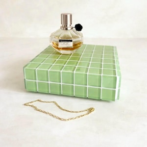 May include: A clear perfume bottle with a silver cap and black flower accent sits atop a light green tiled block. A gold chain necklace rests in front of the block, creating a minimalist aesthetic.