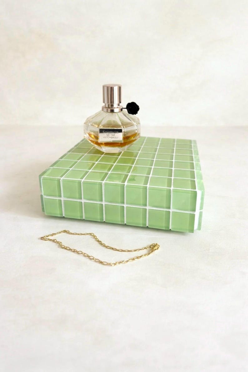May include: A clear perfume bottle with a silver cap and black flower accent sits atop a light green tiled block. A gold chain necklace rests in front of the block, creating a minimalist aesthetic.