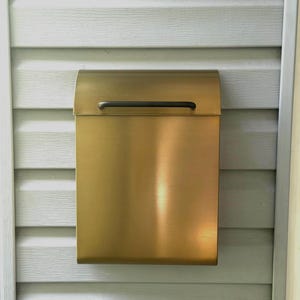 May include: A rectangular wall-mounted mailbox with a brushed brass finish and a smooth, curved top lid. The mailbox features a thin, horizontal black handle centered on the front flap for opening, providing a clean and functional design.