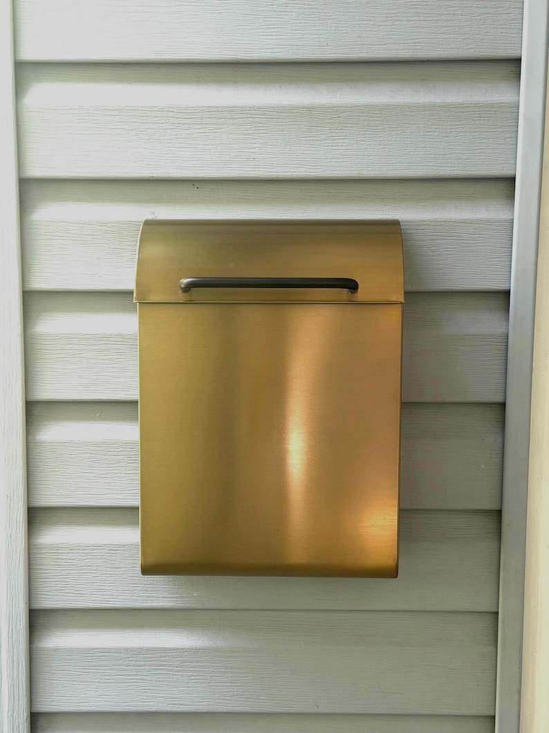 May include: A rectangular wall-mounted mailbox with a brushed brass finish and a smooth, curved top lid. The mailbox features a thin, horizontal black handle centered on the front flap for opening, providing a clean and functional design.