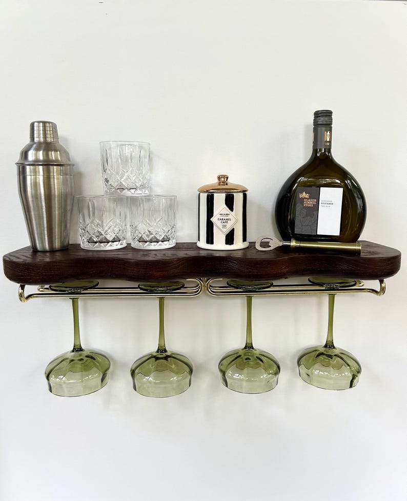 May include: A dark wood wall-mounted shelf with a live edge, featuring a gold metal rack holding four green-tinted wine glasses. On the shelf are a stainless steel cocktail shaker, three crystal tumblers, a striped jar, a bottle, and a bottle opener.