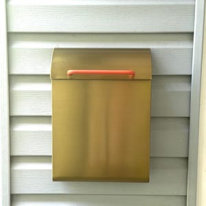 May include: A rectangular wall-mounted mailbox with a brushed gold metallic finish and a horizontal coral-colored handle attached to the top lid. The mailbox features a smooth, clean design with a curved top flap and a flat front panel.