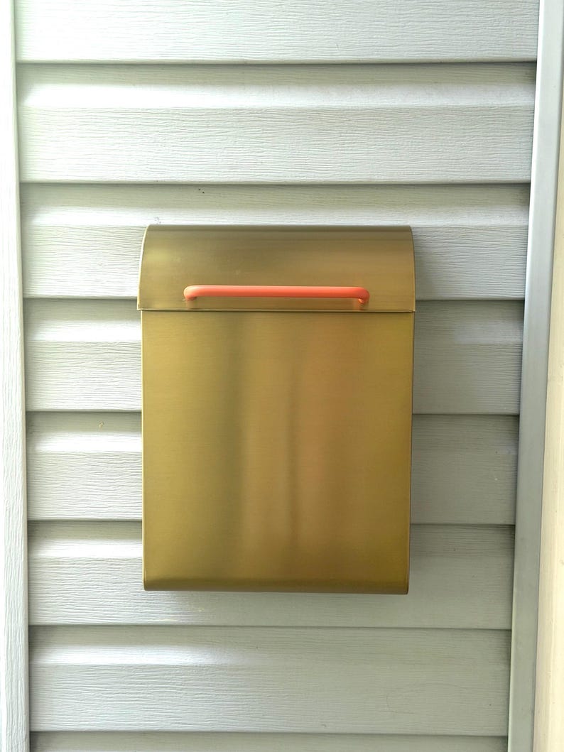 May include: A rectangular wall-mounted mailbox with a brushed gold metallic finish and a horizontal coral-colored handle attached to the top lid. The mailbox features a smooth, clean design with a curved top flap and a flat front panel.