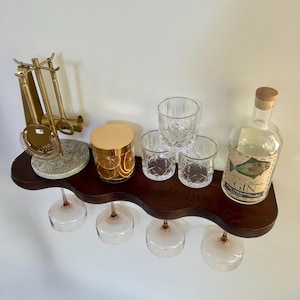 May include: A dark wood wavy shelf holding a gold bar tool set on a stone base, a glass jar with dried citrus slices, three crystal tumblers, and a clear glass bottle labeled 50 Fathoms GIN with a fish illustration, with four wine glasses hanging below.