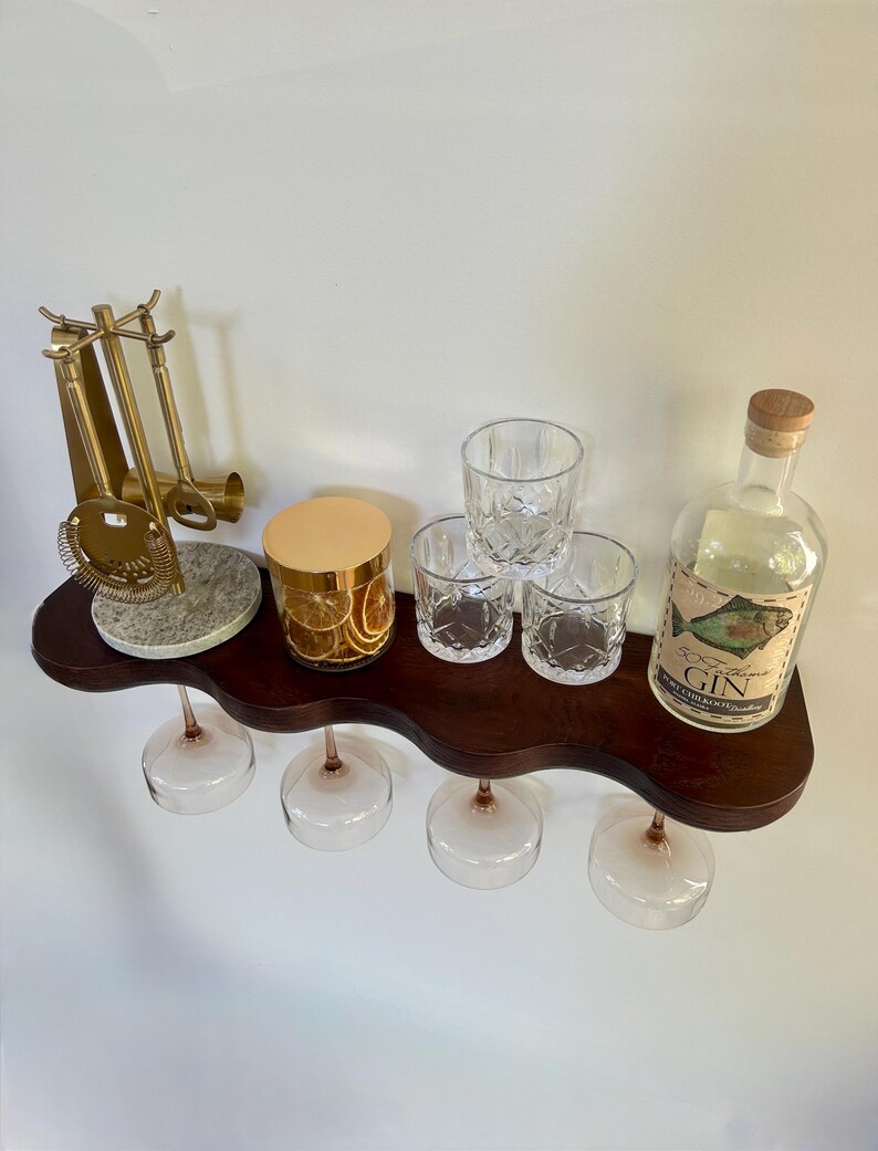 May include: A dark wood wavy shelf holding a gold bar tool set on a stone base, a glass jar with dried citrus slices, three crystal tumblers, and a clear glass bottle labeled 50 Fathoms GIN with a fish illustration, with four wine glasses hanging below.