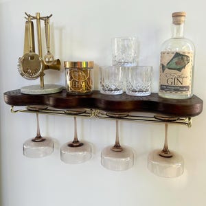 May include: A dark wood wall-mounted shelf with a gold metal rack holding four stemmed glasses. On top sits a gold bar tool stand, a jar labeled DEHY, three crystal tumblers, and a bottle of 50 Fathoms Gin with a fish illustration on the label.