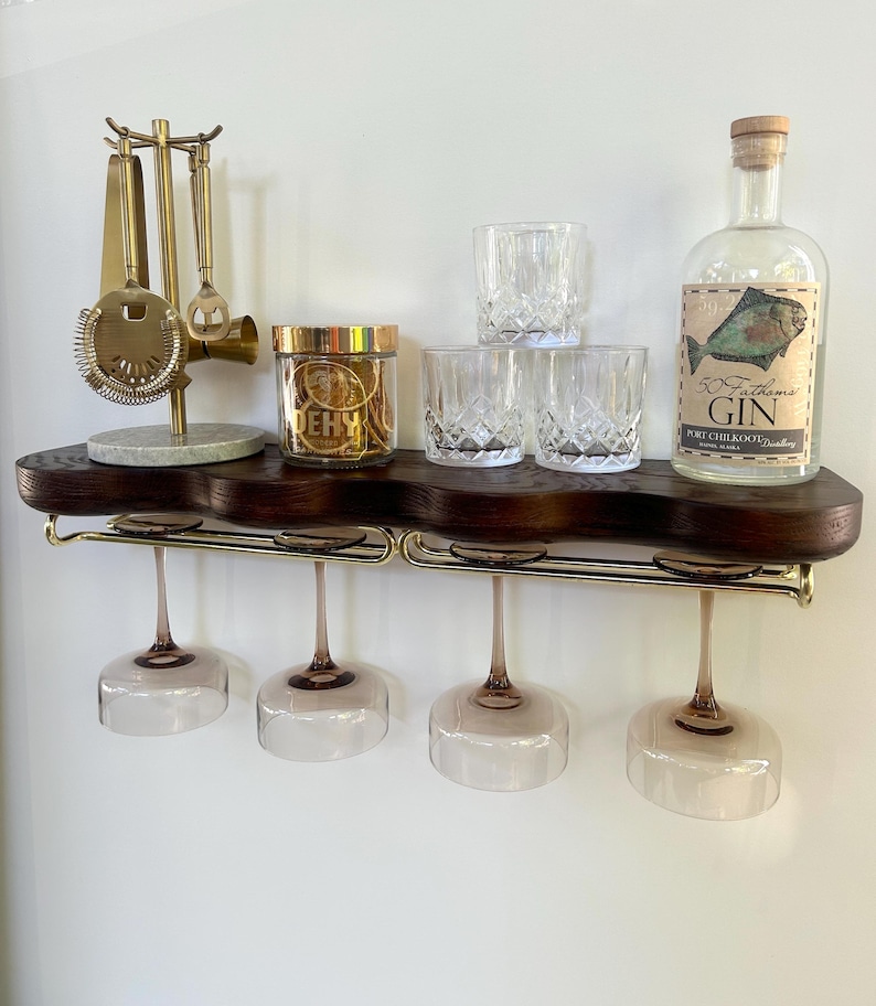 May include: A dark wood wall-mounted shelf with a gold metal rack holding four stemmed glasses. On top sits a gold bar tool stand, a jar labeled DEHY, three crystal tumblers, and a bottle of 50 Fathoms Gin with a fish illustration on the label.