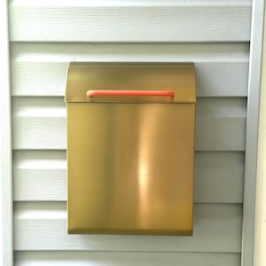 May include: A rectangular wall-mounted mailbox with a brushed gold metallic finish. The mailbox features a curved top lid and a horizontal handle in a bright orange color, providing a contrast to the metallic body of the mailbox unit.