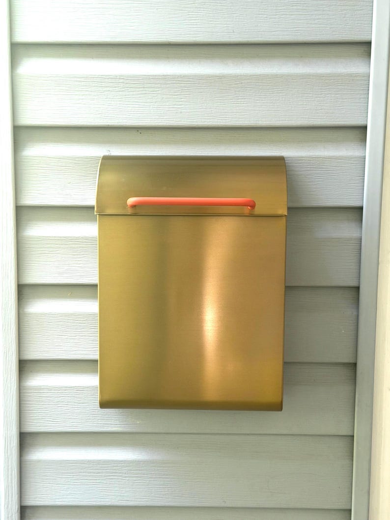 May include: A rectangular wall-mounted mailbox with a brushed gold metallic finish. The mailbox features a curved top lid and a horizontal handle in a bright orange color, providing a contrast to the metallic body of the mailbox unit.