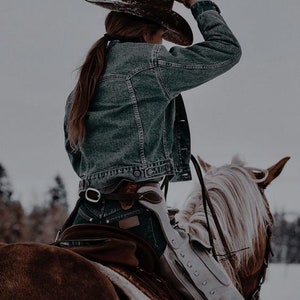 May include: A woman wearing a denim jacket and a brown cowboy hat rides a brown and white horse. The horse is wearing a saddle and a white leather chaps.