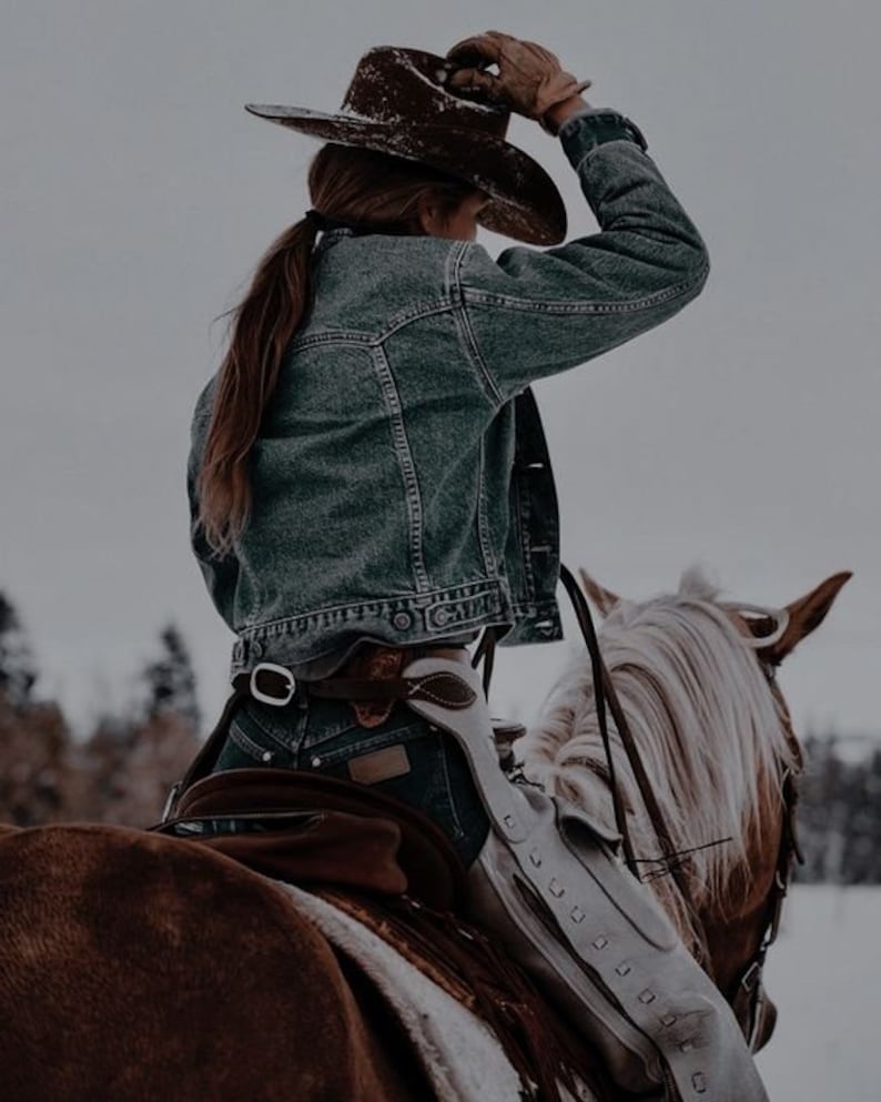 May include: A woman wearing a denim jacket and a brown cowboy hat rides a brown and white horse. The horse is wearing a saddle and a white leather chaps.