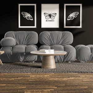 May include: A gray modular sofa with a gray ottoman and a round wooden coffee table. The sofa and ottoman are covered in a soft, plush fabric. The coffee table has a white marble top and a wooden base. Three black and white framed prints of butterflies are hanging on the wall behind the sofa. The prints have the word "Butterfly" written on them. A green plant is in a pot on a stand to the right of the sofa.