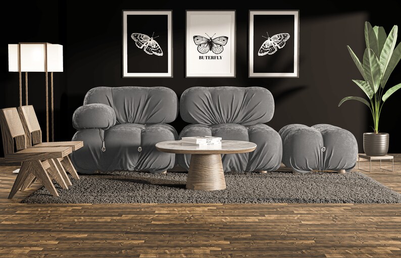 May include: A gray modular sofa with a gray ottoman and a round wooden coffee table. The sofa and ottoman are covered in a soft, plush fabric. The coffee table has a white marble top and a wooden base. Three black and white framed prints of butterflies are hanging on the wall behind the sofa. The prints have the word "Butterfly" written on them. A green plant is in a pot on a stand to the right of the sofa.