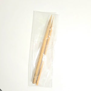 May include: Two wooden skewers in a clear plastic bag. The skewers are approximately 15 cm long and have a pointed tip.