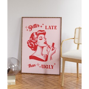 Pu&ograve; includere: Una stampa incorniciata rosa con un'illustrazione rossa di una donna che si applica il rossetto. Il testo sulla stampa recita "Better Late than Ugly."