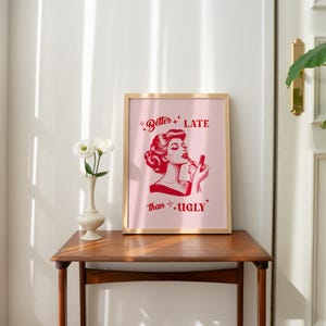 Pu&ograve; includere: Una stampa incorniciata con uno sfondo rosa e testo rosso che recita "Better Late Than Ugly". Al centro della stampa &egrave; presente un'illustrazione rossa di una donna che si applica il rossetto.