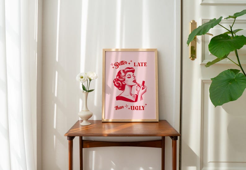 Pu&ograve; includere: Una stampa incorniciata con uno sfondo rosa e testo rosso che recita "Better Late Than Ugly". Al centro della stampa &egrave; presente un'illustrazione rossa di una donna che si applica il rossetto.