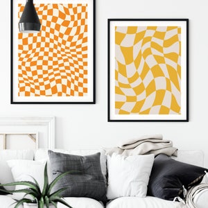 May include: Two abstract art prints with a checkered pattern in shades of orange and yellow. The prints are framed in black and hung on a white wall above a white couch.