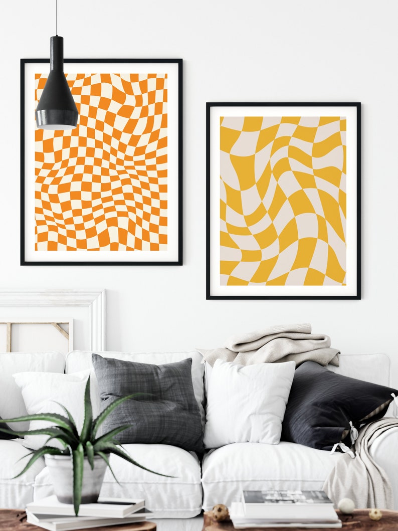 May include: Two abstract art prints with a checkered pattern in shades of orange and yellow. The prints are framed in black and hung on a white wall above a white couch.