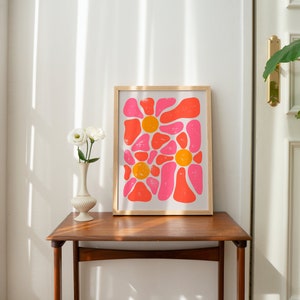 May include: A framed print of an abstract floral design with pink and orange shapes. The print is on a white background and is framed in a light wood frame.