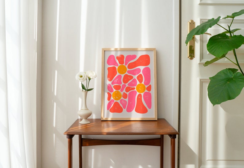 May include: A framed print of an abstract floral design with pink and orange shapes. The print is on a white background and is framed in a light wood frame.