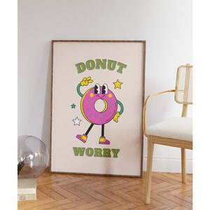 May include: A framed print with a pink donut character with green eyes and a yellow smile. The donut is wearing green shoes and has a green peace sign on its hand. The text "DONUT WORRY" is written in green letters on a beige background.