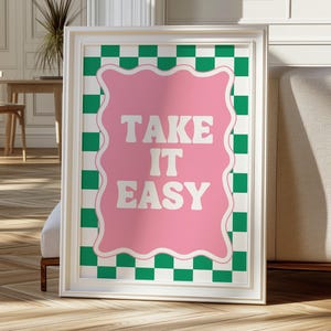 May include: A white framed print with a pink background and green and white checkered border. The text "TAKE IT EASY" is written in white on the pink background.
