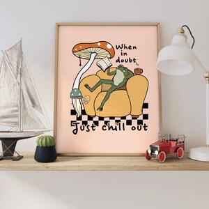 May include: A pink poster with a cartoon frog sitting in a yellow armchair, reading a book and holding a cup. The frog is under a large red and white mushroom. The text on the poster reads "When in doubt, just chill out".
