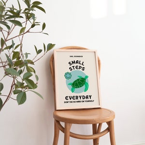 Pu&ograve; includere: Un poster motivazionale incorniciato con uno sfondo blu chiaro e un'illustrazione di una tartaruga verde. Il testo recita "Promemoria quotidiano: Piccoli passi ogni giorno. Non essere troppo duro con te stesso."