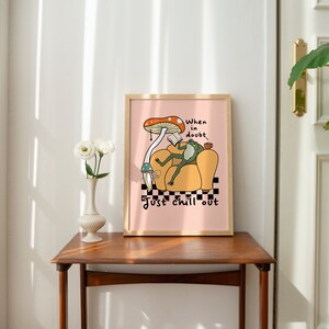 May include: A framed print with a pink background featuring a cartoon frog sitting in a yellow armchair. The frog is wearing a green shirt and holding a book. A mushroom with a red cap is above the frog. The text "When in doubt, just chill out" is written below the frog.
