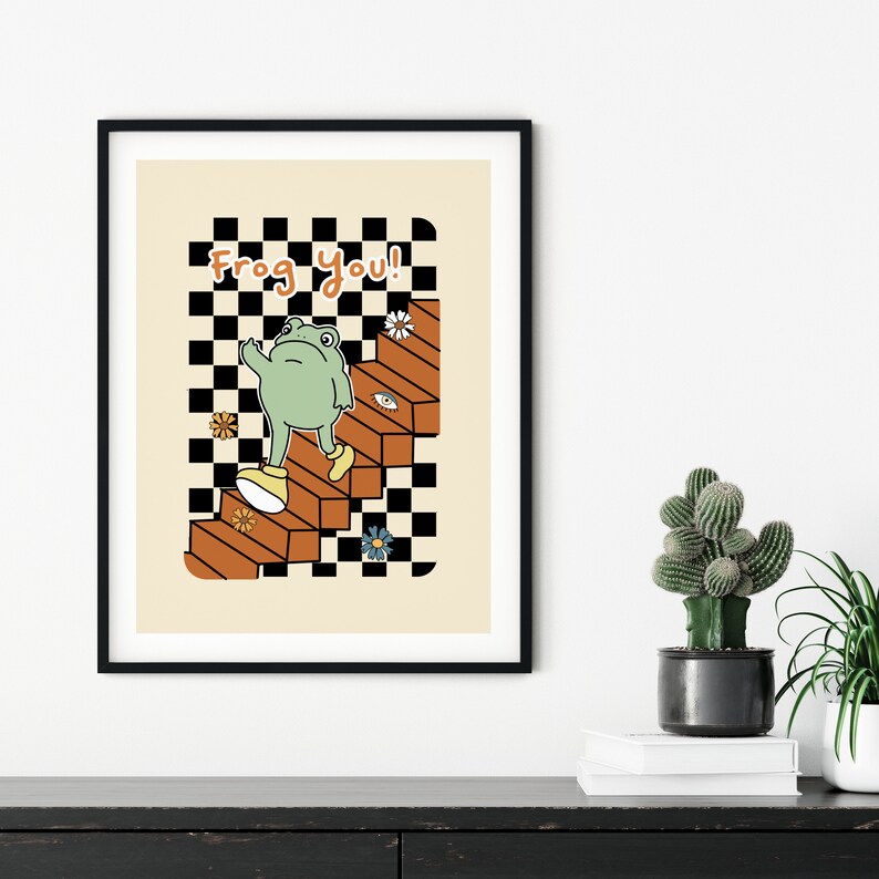 May include: A black framed print with a light beige background featuring a green frog wearing yellow shoes walking up a set of orange stairs with a black and white checkered pattern. The text "Frog You!" is written in black at the top of the print.