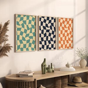 May include: Three framed abstract prints with a checkered pattern. The prints are in shades of blue, green, and orange. The prints are hanging on a white wall above a wooden table.